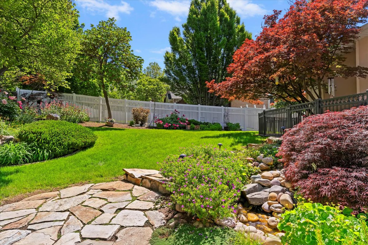 Landscaped backyard featuring a stone pathway, a manicured lawn, a white picket fence, a tiered rock garden with a water feature, and mature trees including a vibrant Japanese maple
