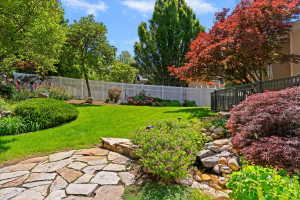 Landscaped backyard featuring a stone pathway, a manicured lawn, a white picket fence, a tiered rock garden with a water feature, and mature trees including a vibrant Japanese maple