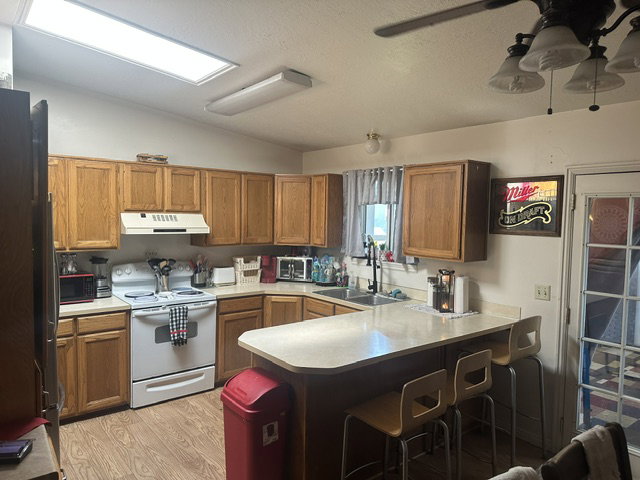 Kitchen featuring wood-finish flooring, wood cabinetry, white appliances, a peninsula with counter seating, and dual basin sink with a high-arc faucet