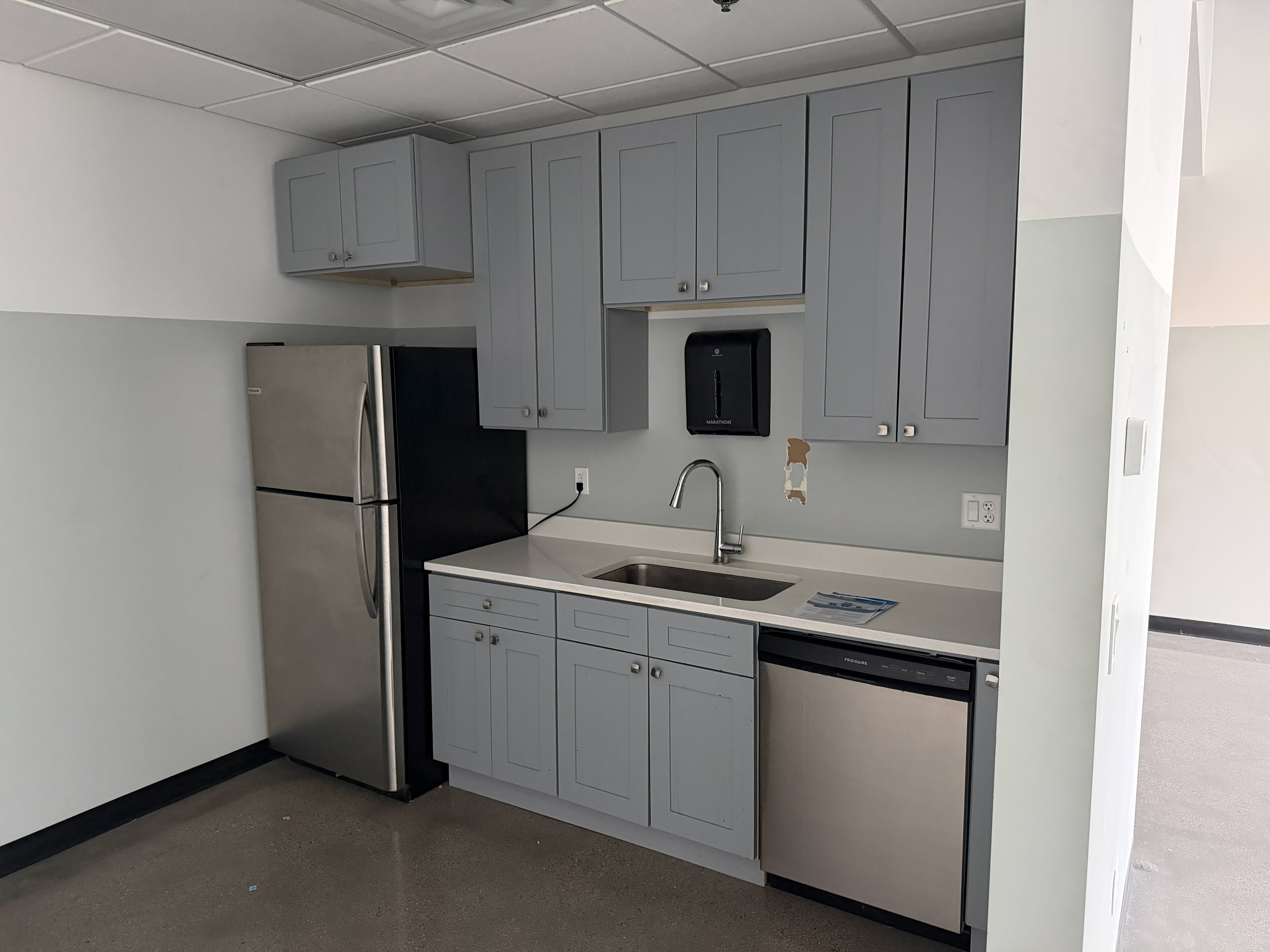 Compact kitchen area featuring gray shaker-style cabinetry, a stainless steel refrigerator, a stainless steel dishwasher, a single-basin stainless steel sink with a chrome faucet, and light-toned countertops