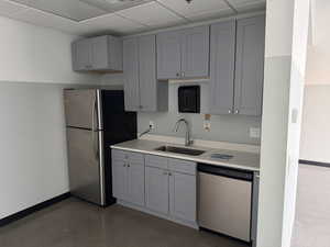 Compact kitchen area featuring gray shaker-style cabinetry, a stainless steel refrigerator, a stainless steel dishwasher, a single-basin stainless steel sink with a chrome faucet, and light-toned countertops