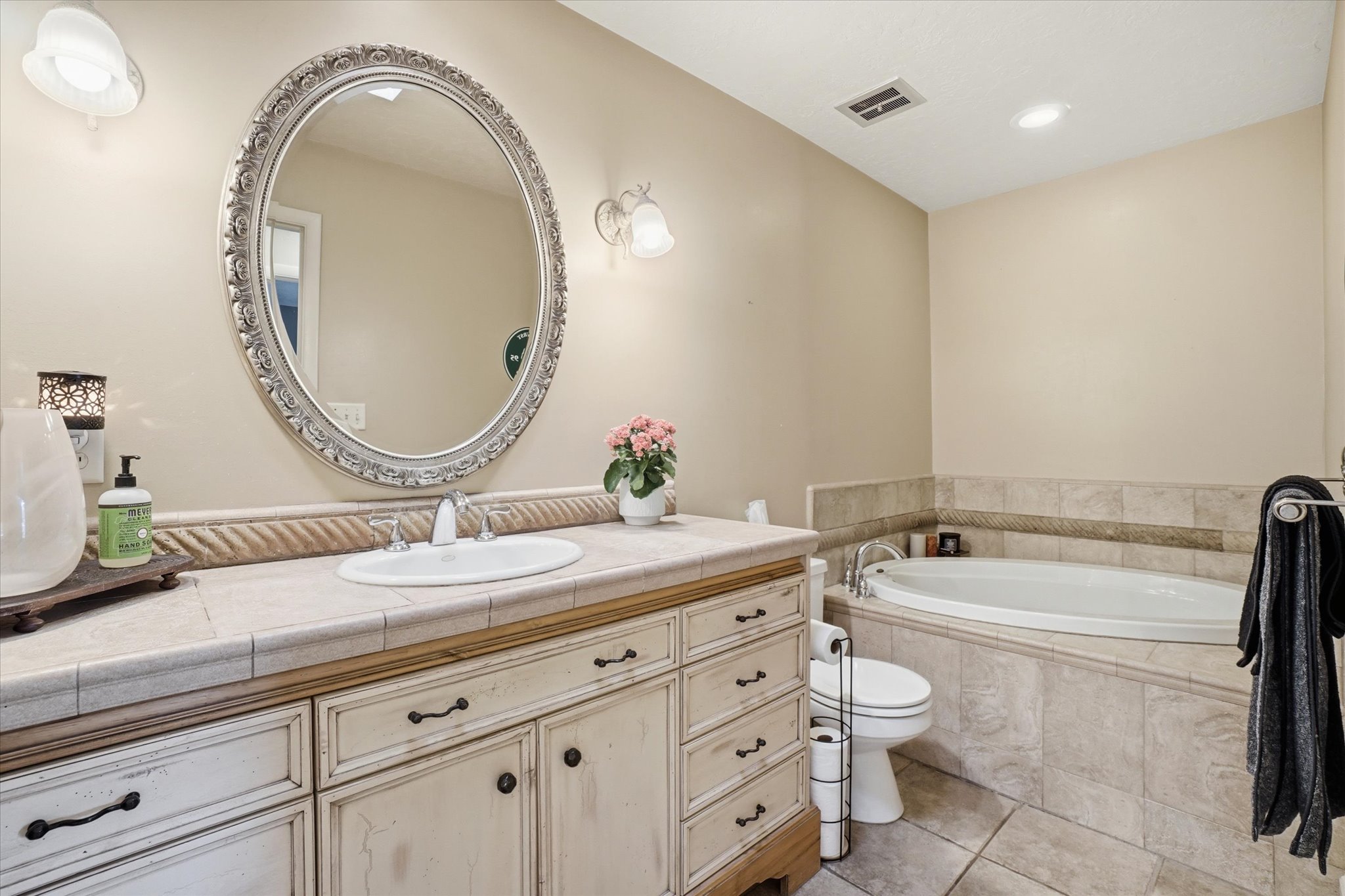 Spacious bathroom featuring a built-in jetted tub, an ornate framed oval mirror, a vanity with distressed cabinetry, a tiled countertop, and recessed lighting