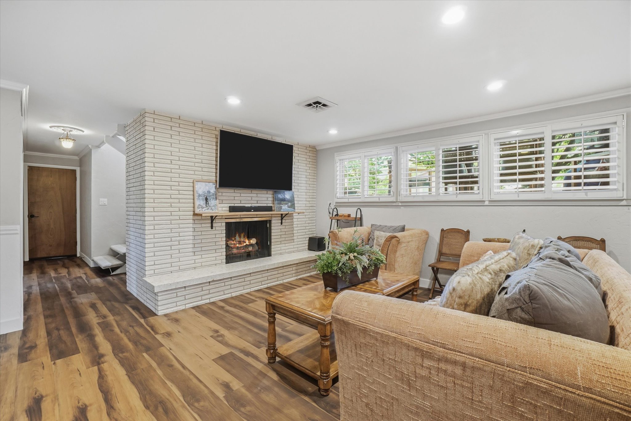 Living area featuring a white brick fireplace with an integrated mantel and hearth, wood-finish flooring, recessed lighting, and multiple windows with plantation shutters