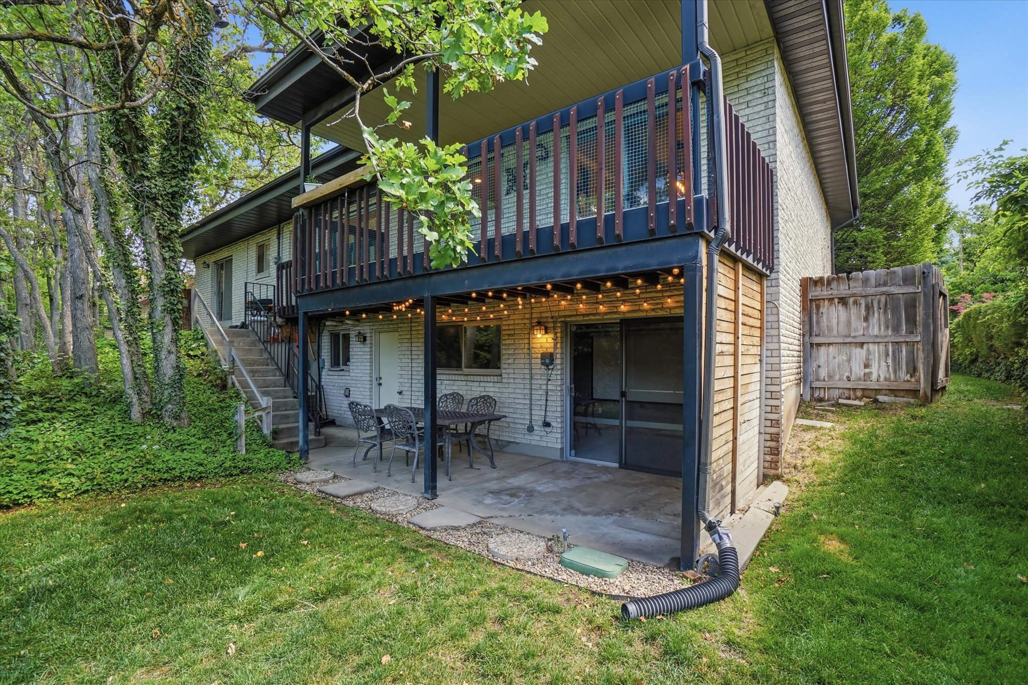 Rear exterior featuring a ground-level patio with overhead string lighting, a second-story balcony with dark railings, and a stairway entry
