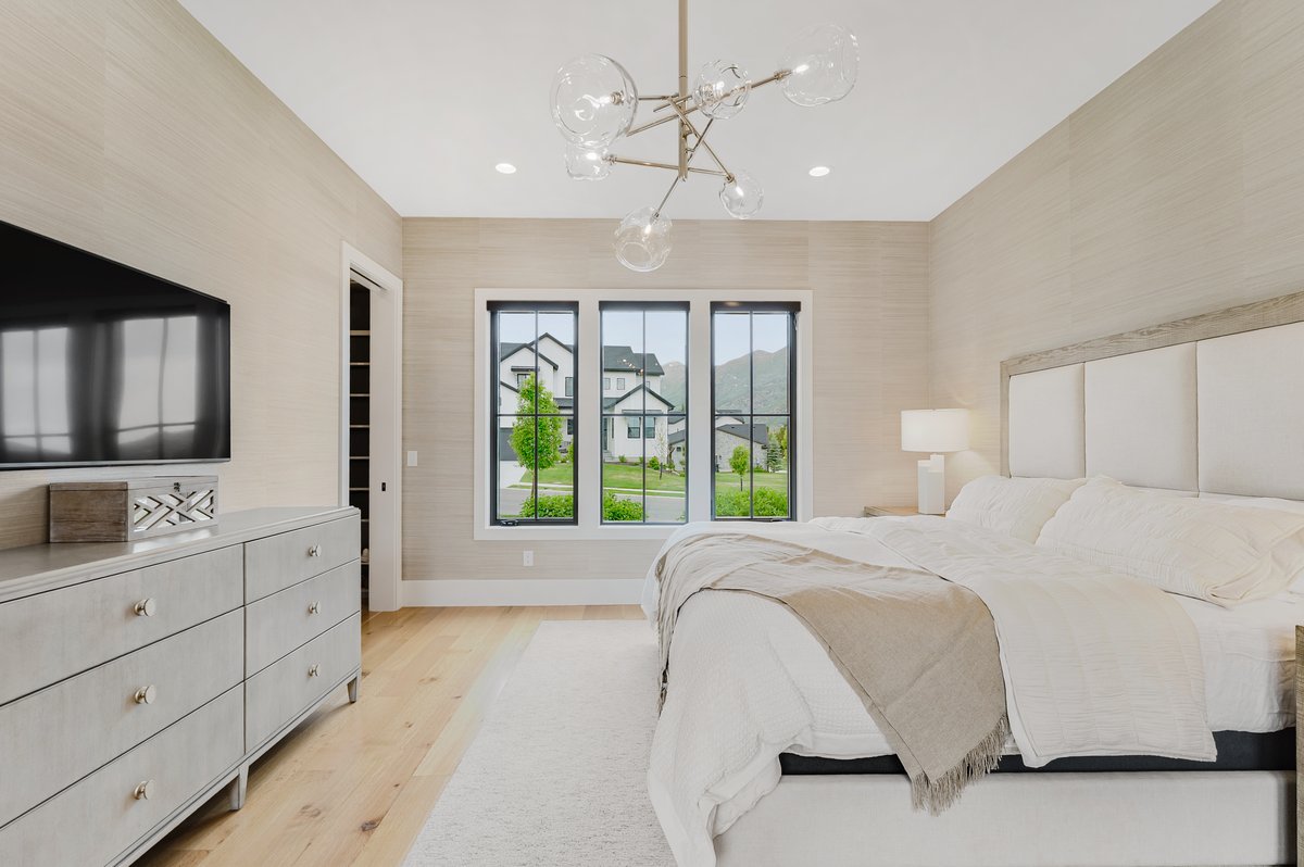 Spacious bedroom featuring light wood-finish flooring, a contemporary chandelier, a large upholstered headboard, and a three-panel window with black frames