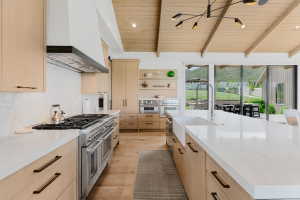 Spacious kitchen featuring a large island with a farmhouse sink, light wood-finish cabinetry, a professional-grade range with a stainless steel hood, and vaulted wood-paneled ceilings