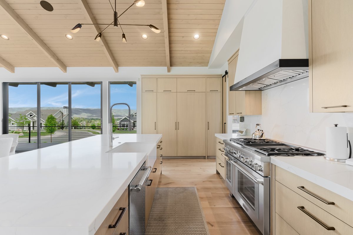 Open-concept kitchen featuring a vaulted wood-paneled ceiling, expansive white countertop island with integrated sink, stainless steel range with overhead hood, wood-finish flooring, and floor-to-ceiling cabinetry