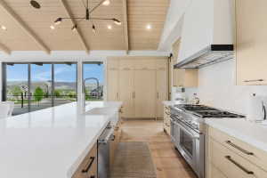 Open-concept kitchen featuring a vaulted wood-paneled ceiling, expansive white countertop island with integrated sink, stainless steel range with overhead hood, wood-finish flooring, and floor-to-ceiling cabinetry