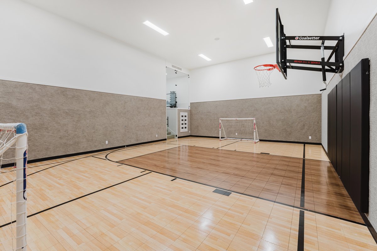 Indoor athletic court featuring hardwood flooring with court markings, an adjustable basketball hoop, and padded wall protection