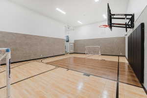 Indoor athletic court featuring hardwood flooring with court markings, an adjustable basketball hoop, and padded wall protection