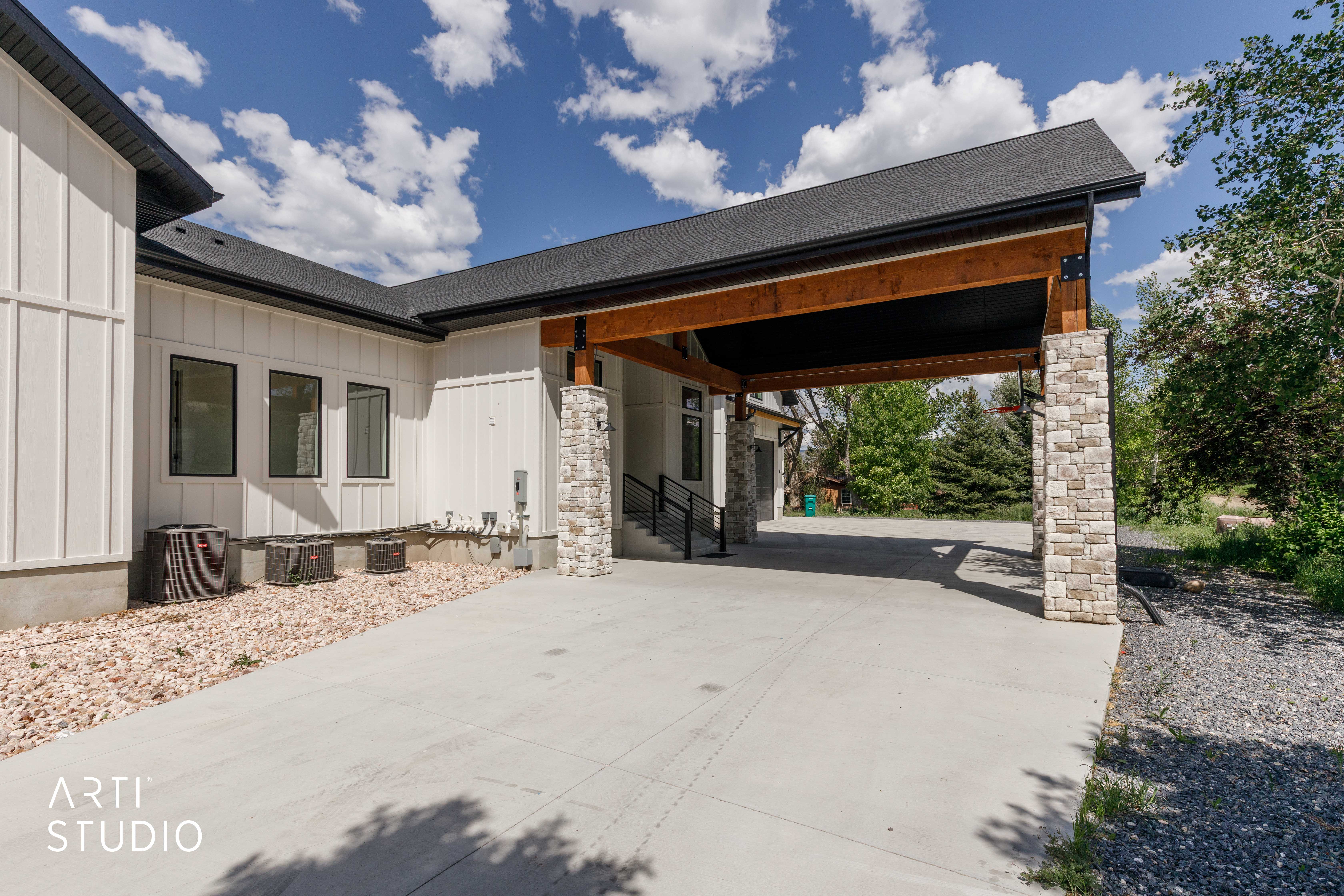 Covered carport featuring exposed wood beams and stacked stone columns, complemented by a board and batten exterior, dark-framed windows, and a gravel landscape bed