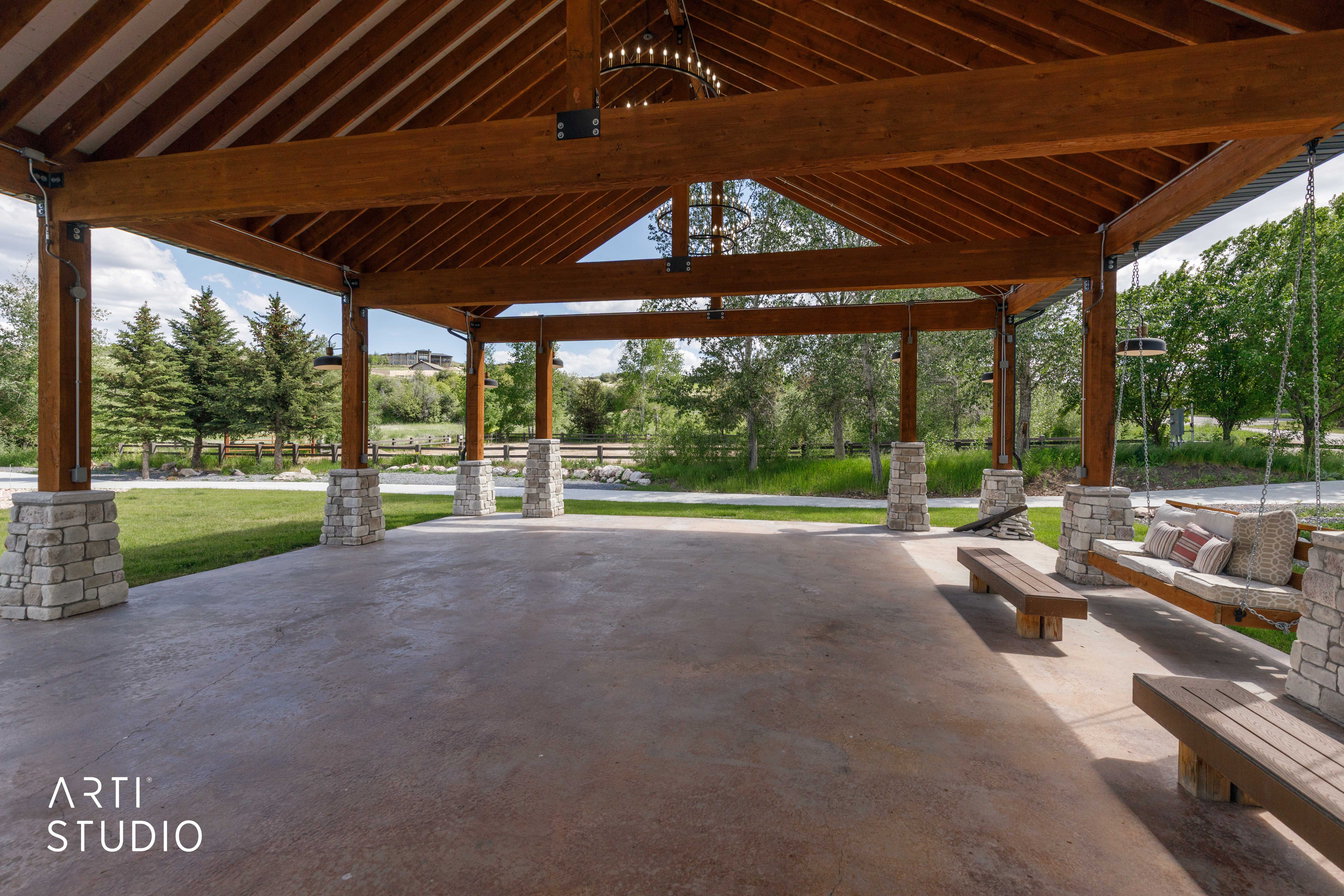 Expansive outdoor pavilion featuring a vaulted wood beam ceiling, stone-clad support columns, a concrete floor, and a suspended chandelier