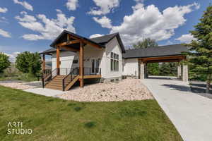 Modern farmhouse exterior featuring white vertical siding, a covered porch with wood posts, a wood-finish deck with black railings, a paved driveway, and an attached carport with stone pillars