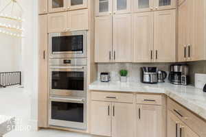 Kitchen featuring light wood-finish cabinetry with modern hardware, integrated stainless steel appliances including a built-in microwave and double oven, marble-look countertops, and a full-height backsplash