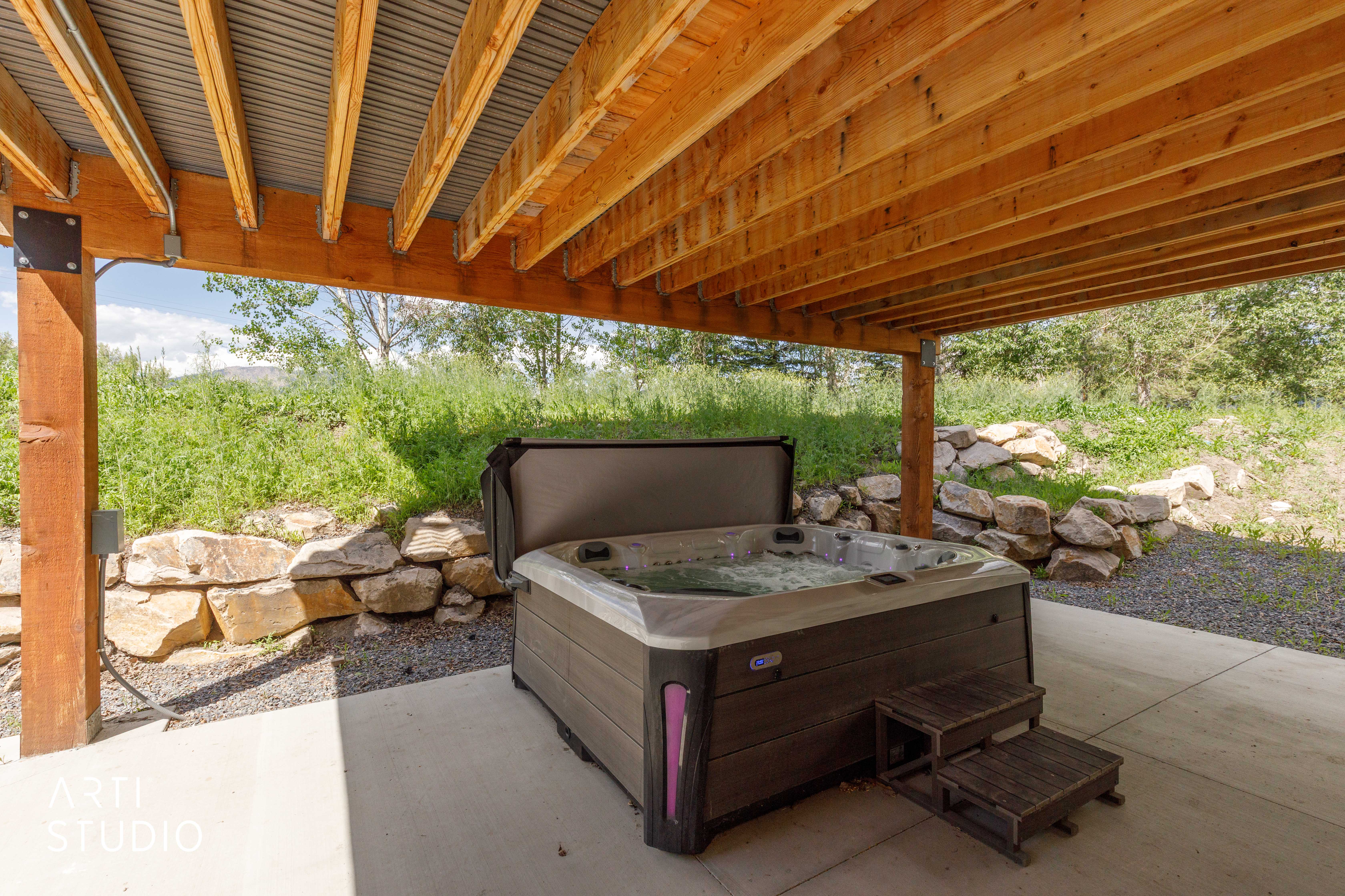 Outdoor spa area featuring a bubbling hot tub with wooden steps, set on a concrete patio under a wooden deck with a corrugated metal ceiling