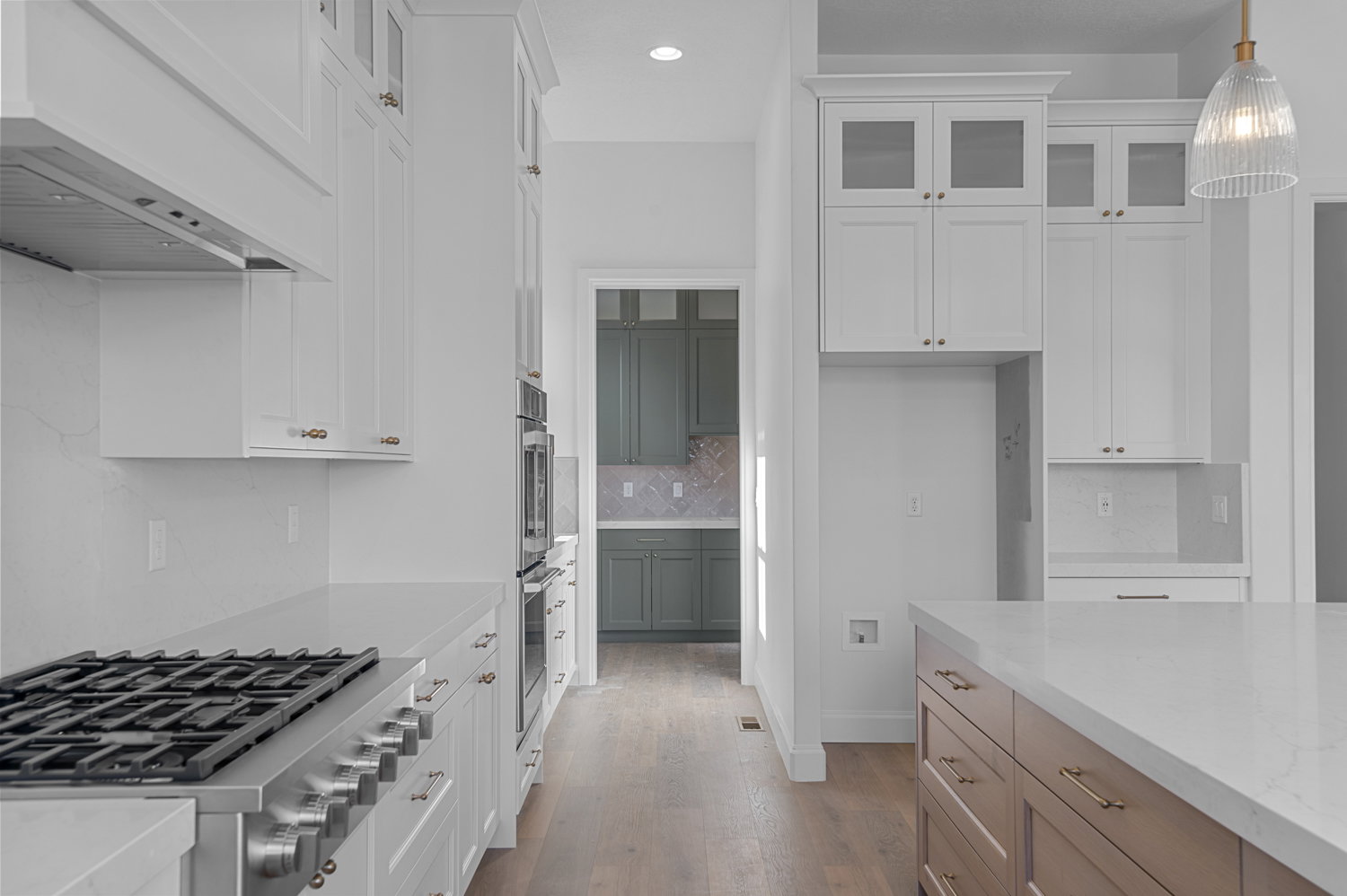 Gourmet kitchen featuring a stainless steel gas range, built-in double ovens, white shaker cabinetry, an expansive island with a light-colored countertop, and wood-finish flooring