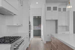 Gourmet kitchen featuring a stainless steel gas range, built-in double ovens, white shaker cabinetry, an expansive island with a light-colored countertop, and wood-finish flooring