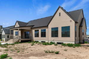 Rear exterior featuring a covered patio with dark railing, multiple large windows, tan horizontal siding, and a dark shingle roof