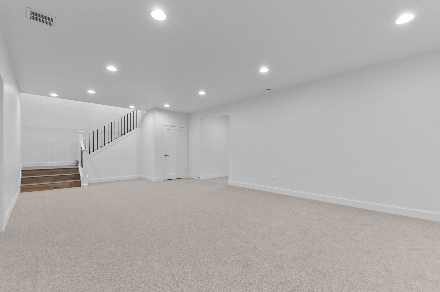 Spacious lower-level room featuring light-toned carpeting, recessed ceiling lighting, a staircase with wood-finish treads and black balusters, and a white interior door