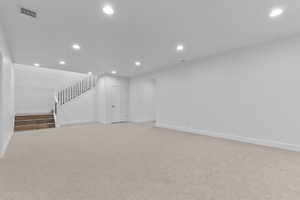 Spacious lower-level room featuring light-toned carpeting, recessed ceiling lighting, a staircase with wood-finish treads and black balusters, and a white interior door