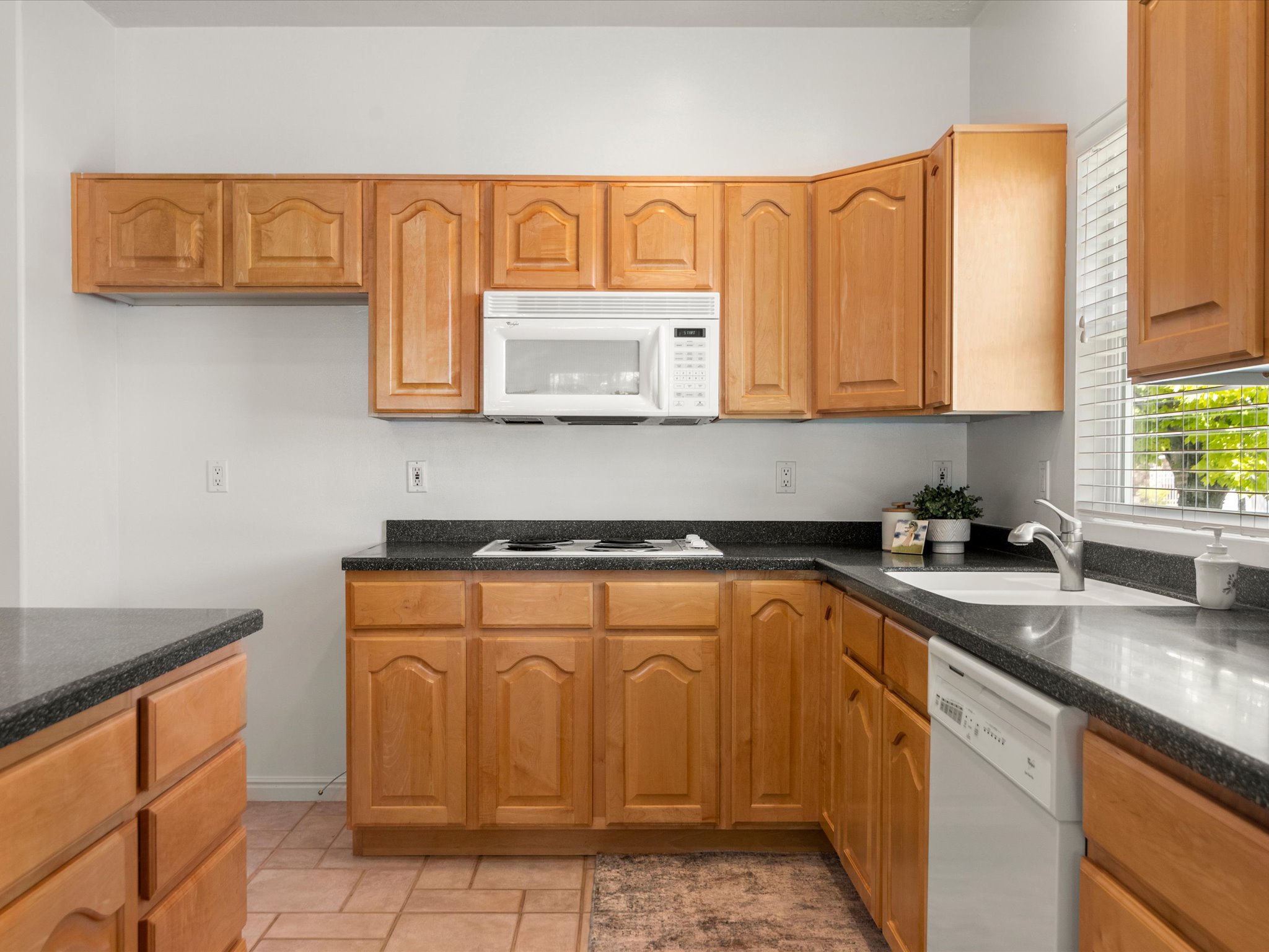 Kitchen featuring light wood-finish cabinetry, dark countertops, a white dishwasher, a white microwave, and a window with blinds