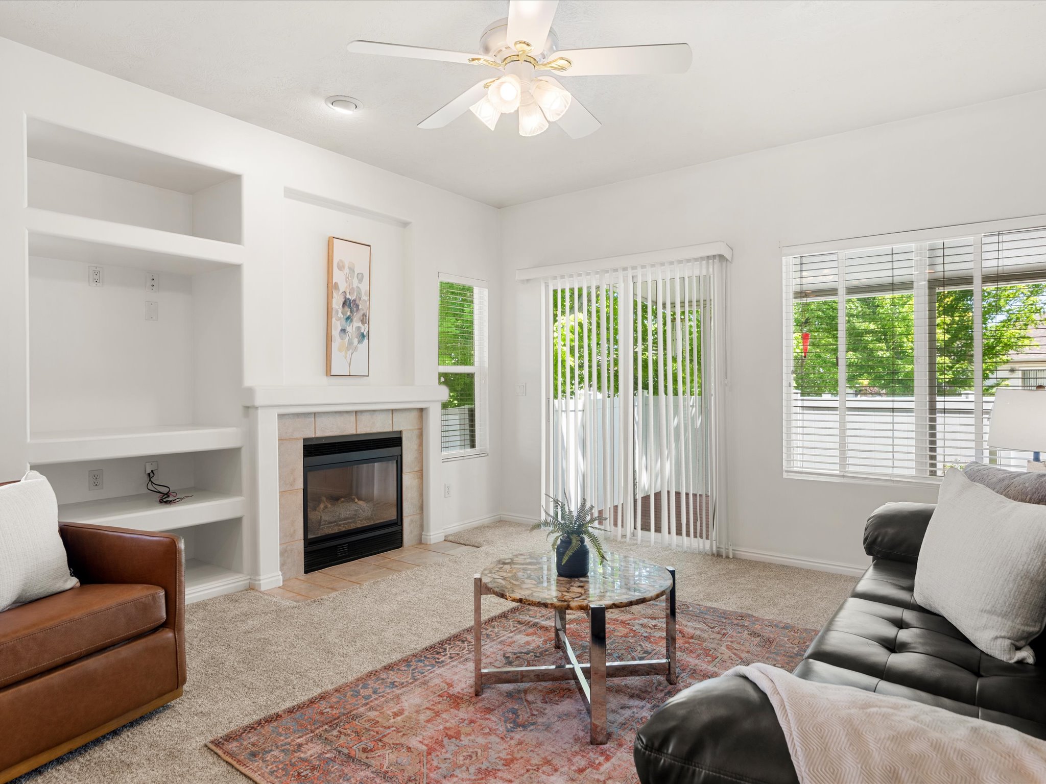Living area featuring a fireplace with a tiled surround, built-in shelving, carpet flooring, and a ceiling fan