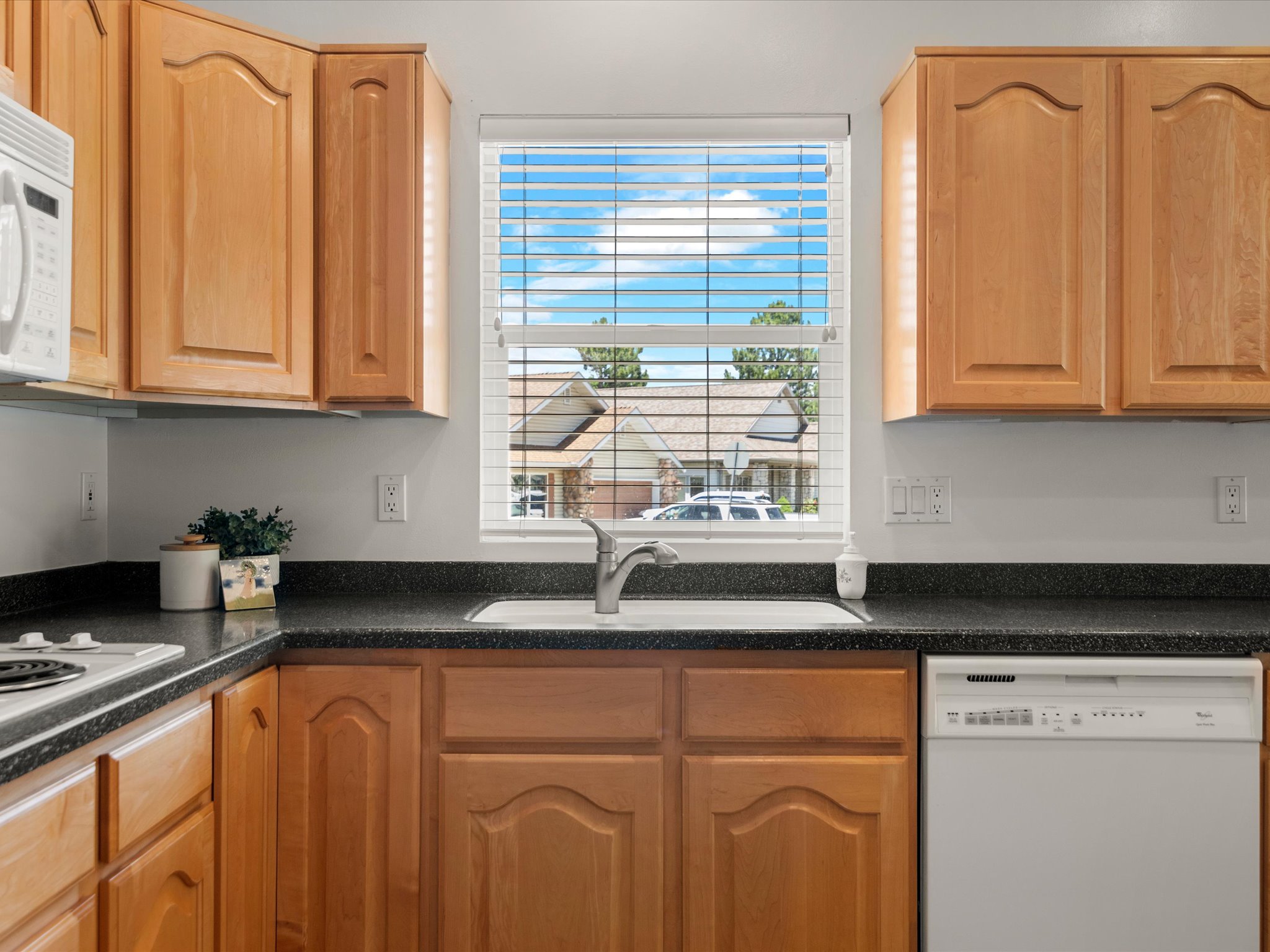 Kitchen featuring light wood cabinetry, a dark countertop, an integrated sink with a stainless steel faucet, and a window with horizontal blinds