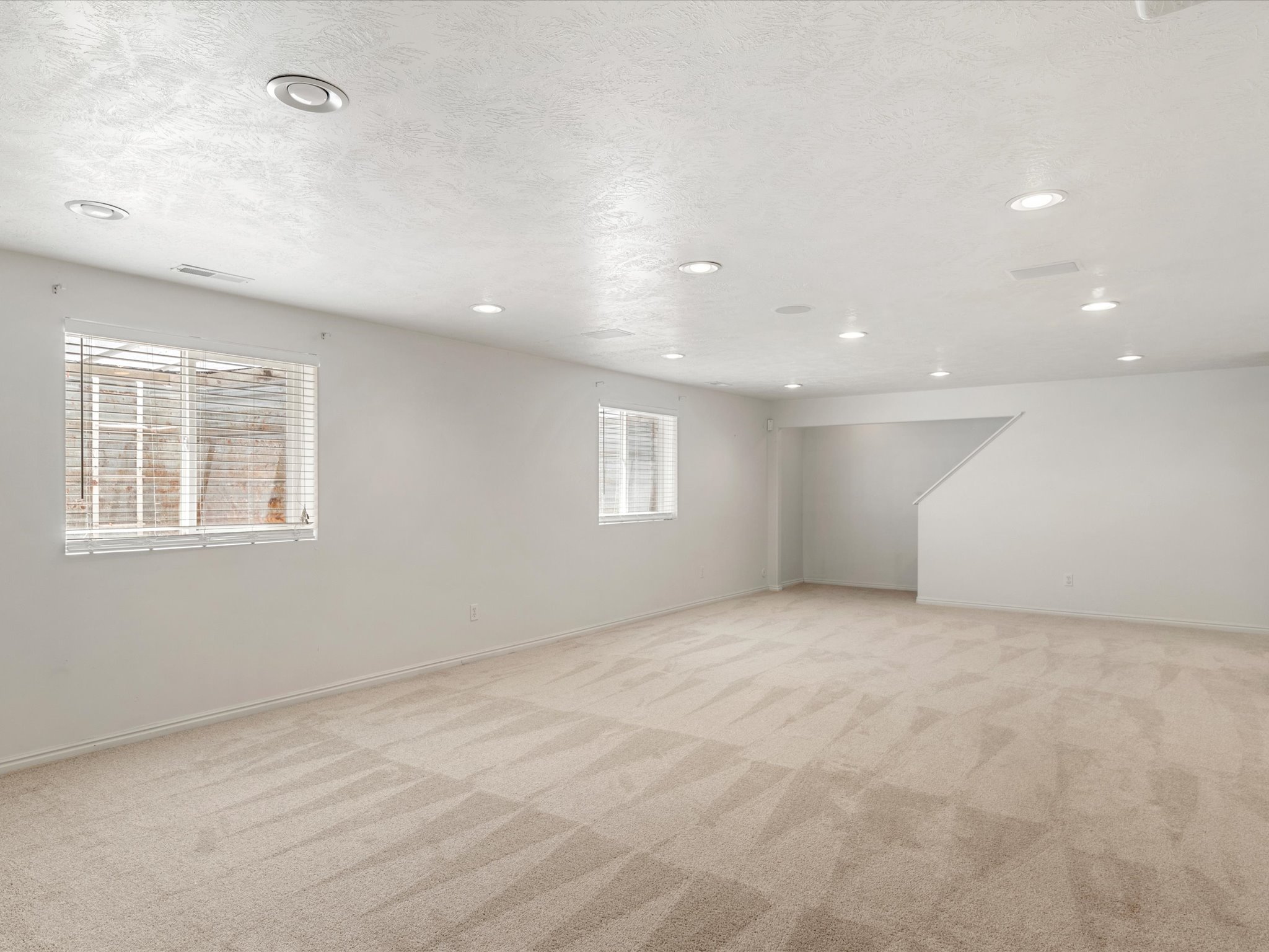 Spacious room featuring neutral carpeting, white walls, multiple windows with blinds, recessed lighting, and a visible staircase
