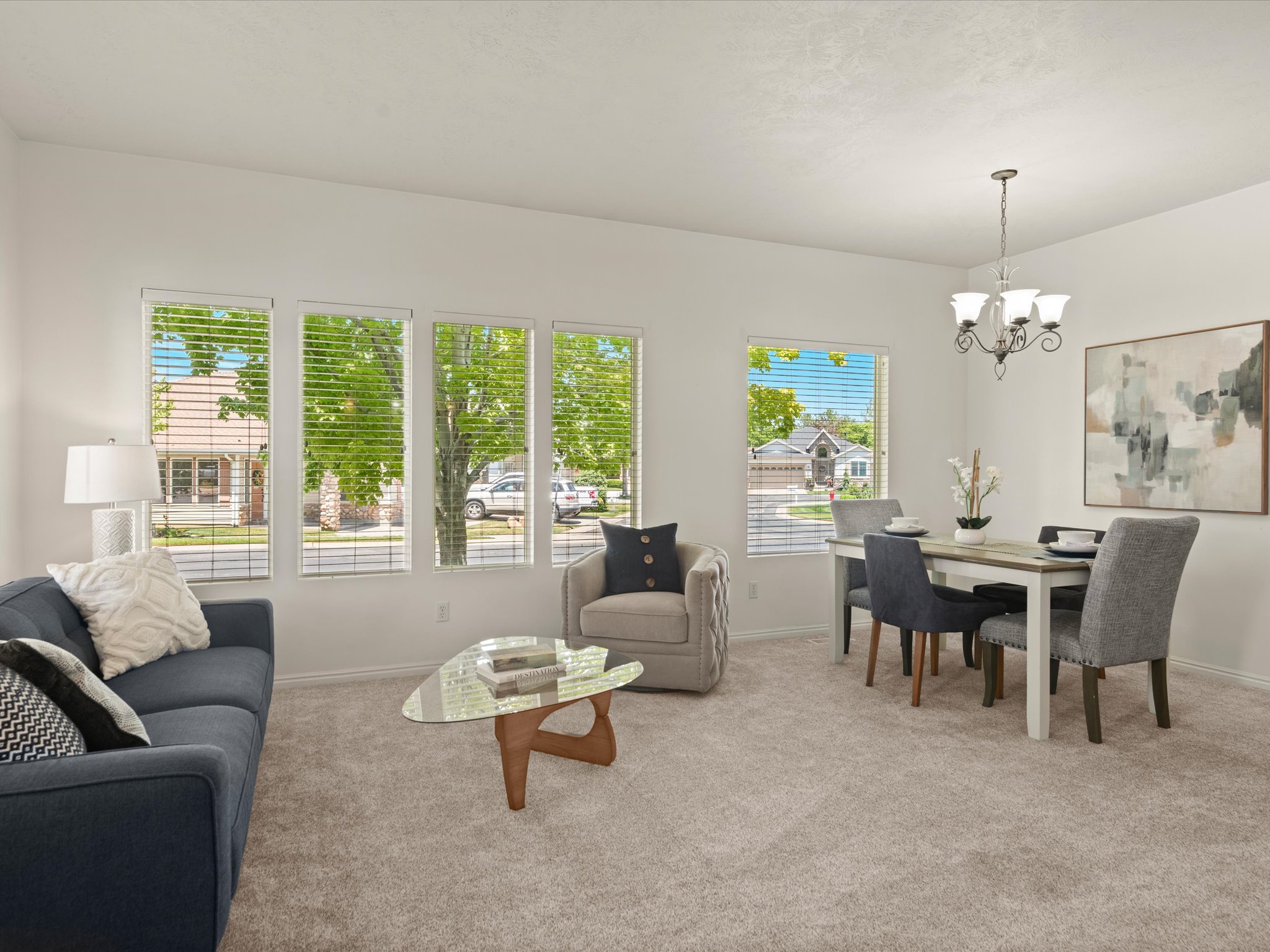 Bright living area featuring multiple large windows with blinds, neutral carpet flooring, and a chandelier over the dining space