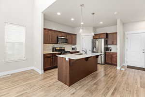 Spacious kitchen featuring rich wood cabinetry, a central island with a sink and white countertop, stainless steel appliances, and a light-toned wood-finish floor