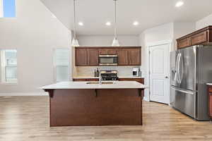 Spacious kitchen featuring a central island with a light-toned countertop, wood-finish flooring, stainless steel appliances, dark wood cabinetry, and a vaulted ceiling with recessed lighting