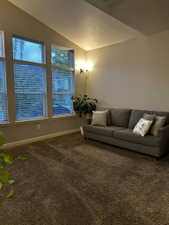 Vaulted ceilings accent a living space featuring multiple windows with blinds, a wall-mounted sconce light fixture, and textured carpet flooring