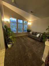 Vaulted ceiling living area featuring a trio of large windows with blinds, sconce lighting, and brown carpeting