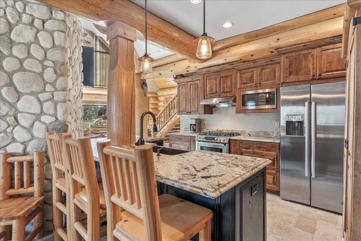 Kitchen featuring stone accent walls, natural wood cabinetry, stainless steel appliances, a multi-level island with a stone countertop, and pendant lighting