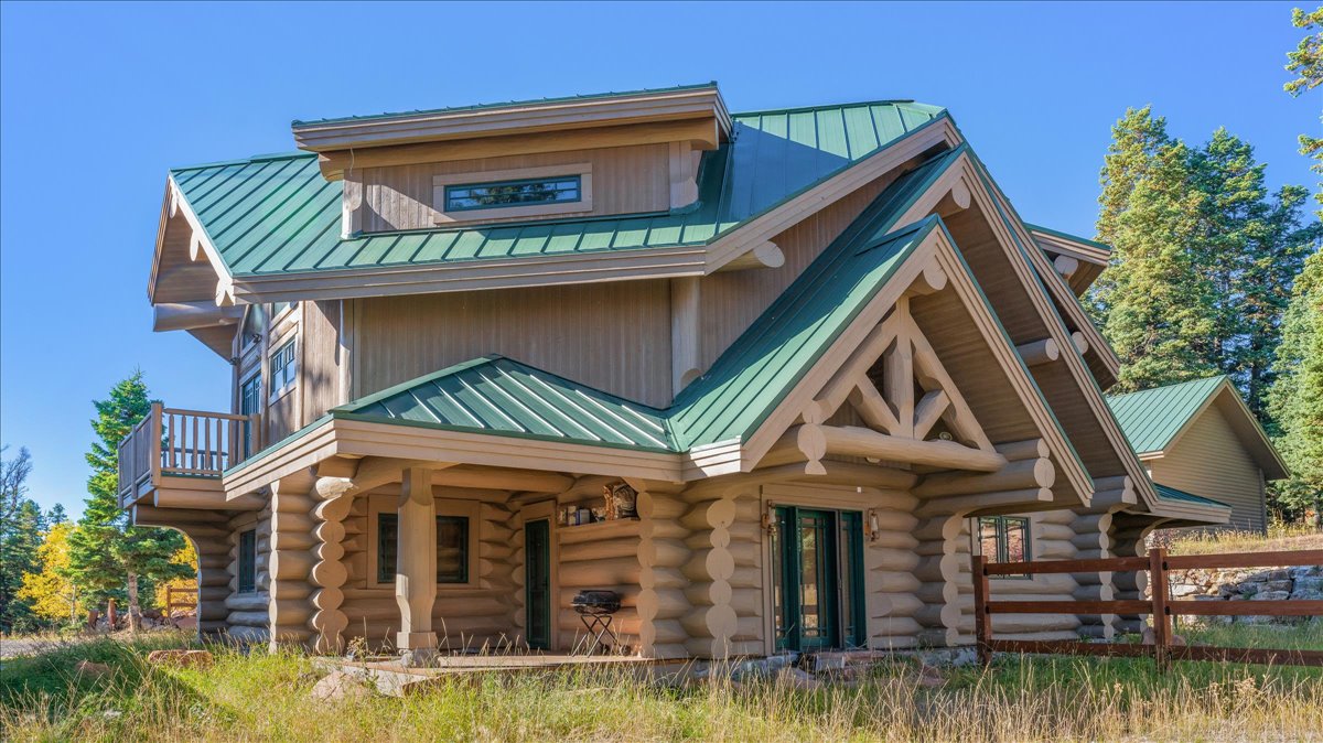 Log-style exterior featuring a green metal roof, a covered front porch with log posts, a second-story balcony with wood railing, prominent gable accents, and a wood fence