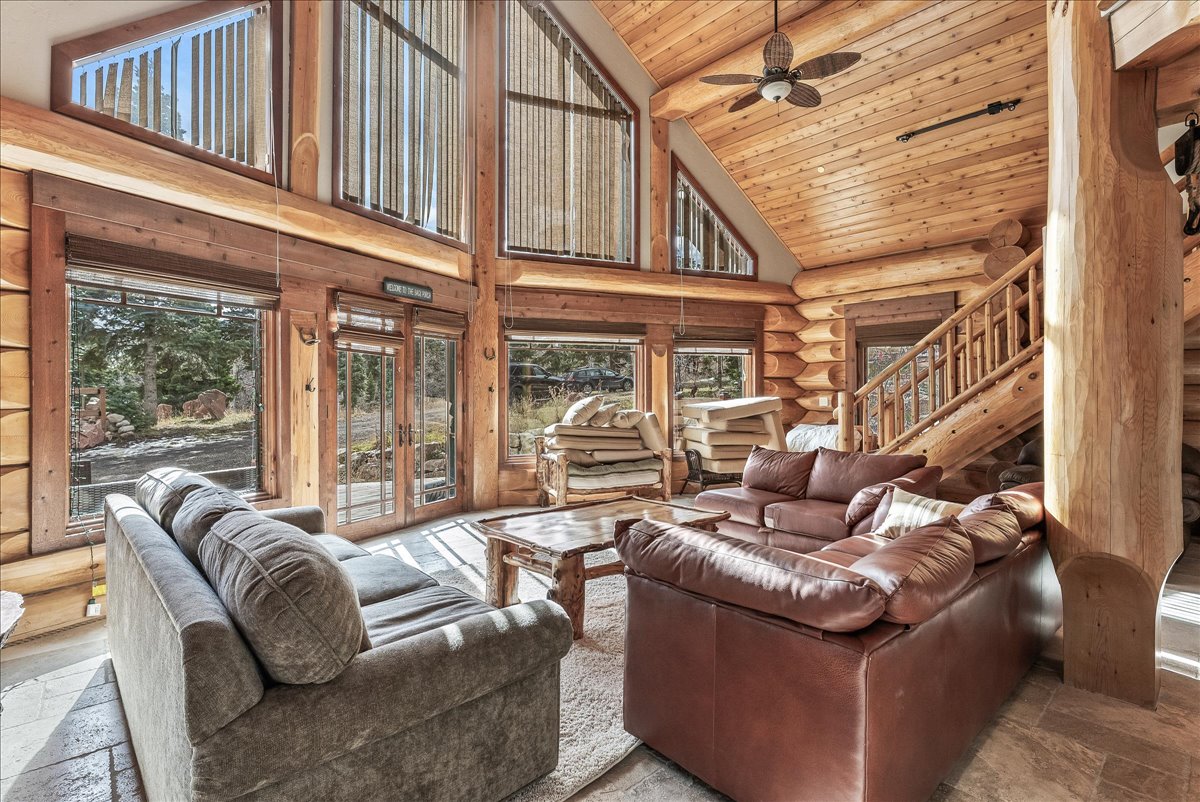 Great room featuring a vaulted wood-paneled ceiling, exposed log beam construction, stone-tile flooring, expansive picture windows, and a wood staircase with balustrade