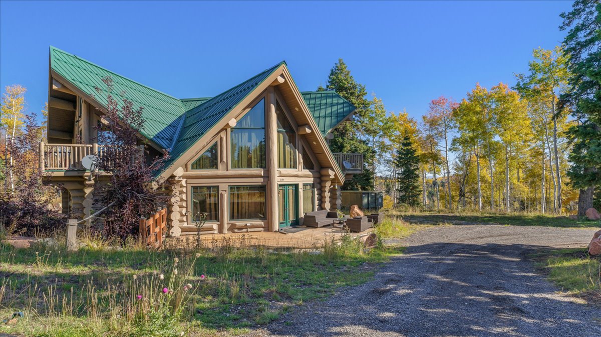 Log cabin exterior featuring a green metal roof, a full-height wall of windows, a balcony with a wood railing, and a gravel driveway