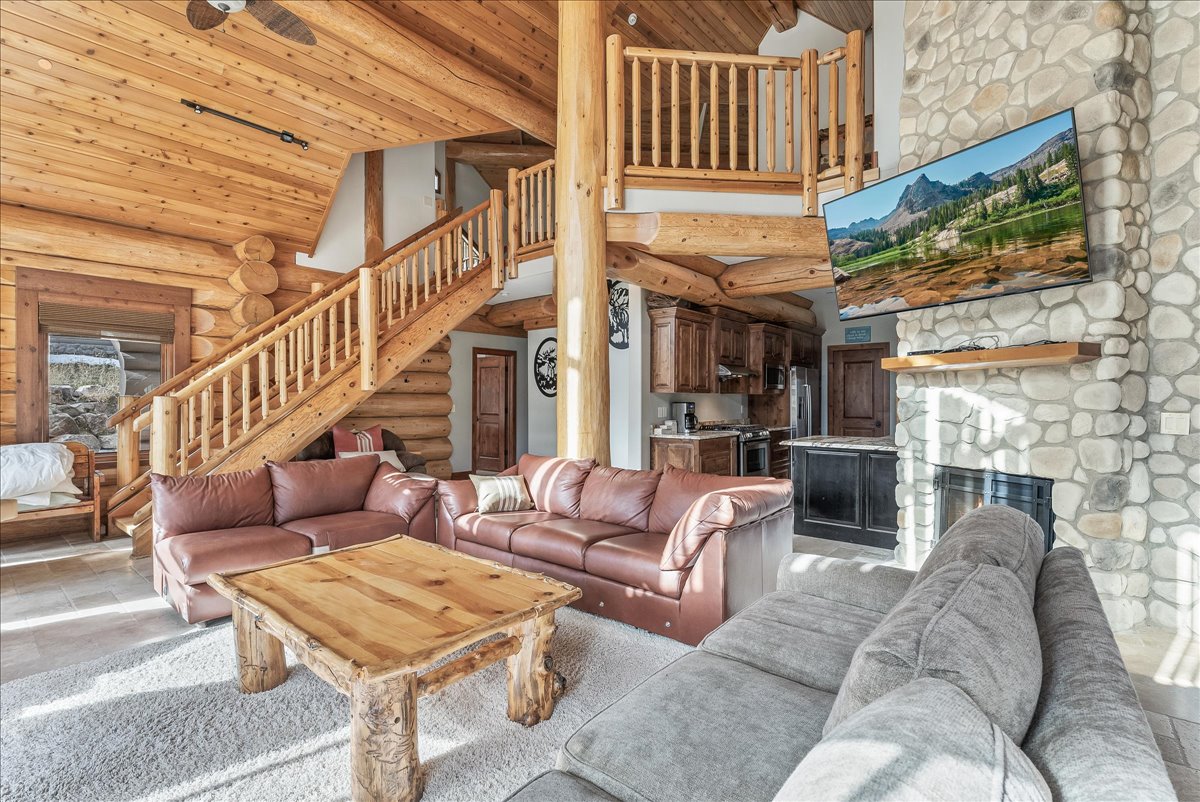Spacious living area featuring log construction, a stone-faced fireplace with a wood mantle, wood-finish flooring, and exposed wood ceiling beams