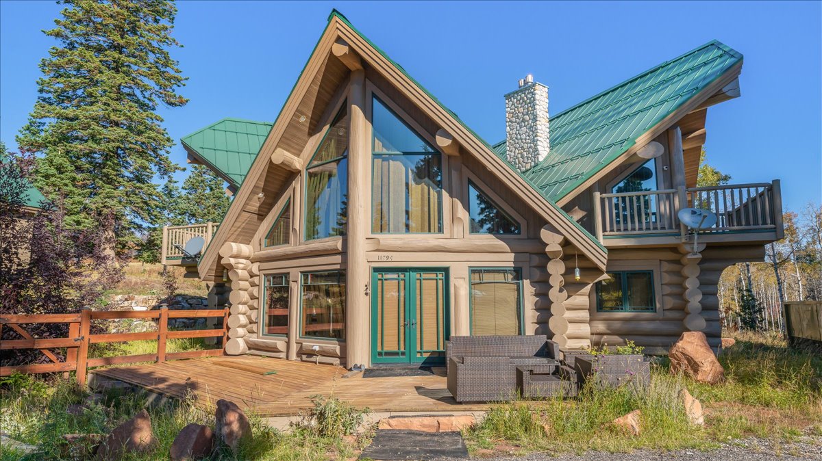 Log cabin exterior featuring a prominent A-frame design, a green metal roof, a stone chimney, extensive window-paneled walls, and a wooden deck