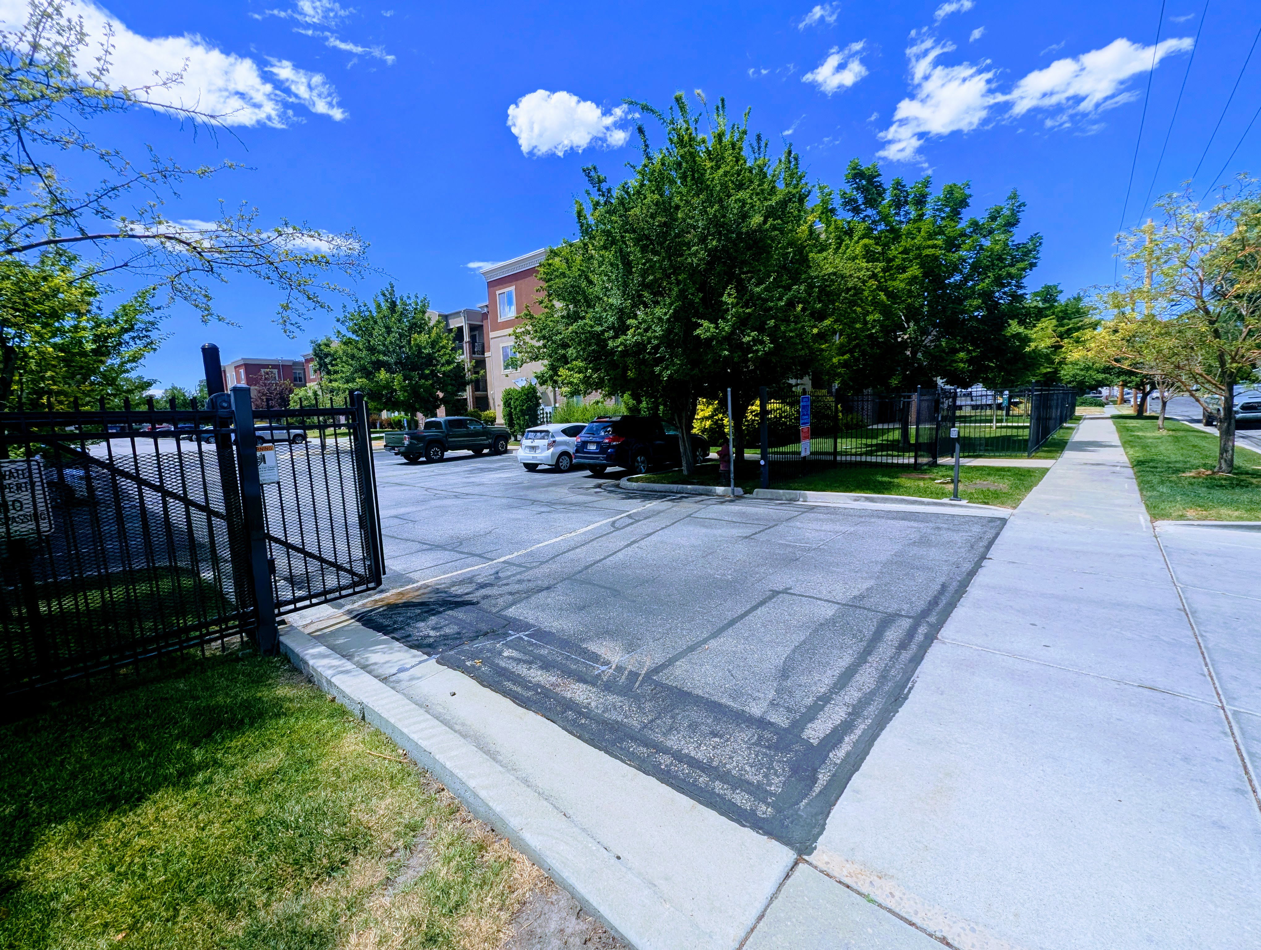 Paved parking area featuring a black metal gate and fence, surrounded by mature trees and a manicured lawn