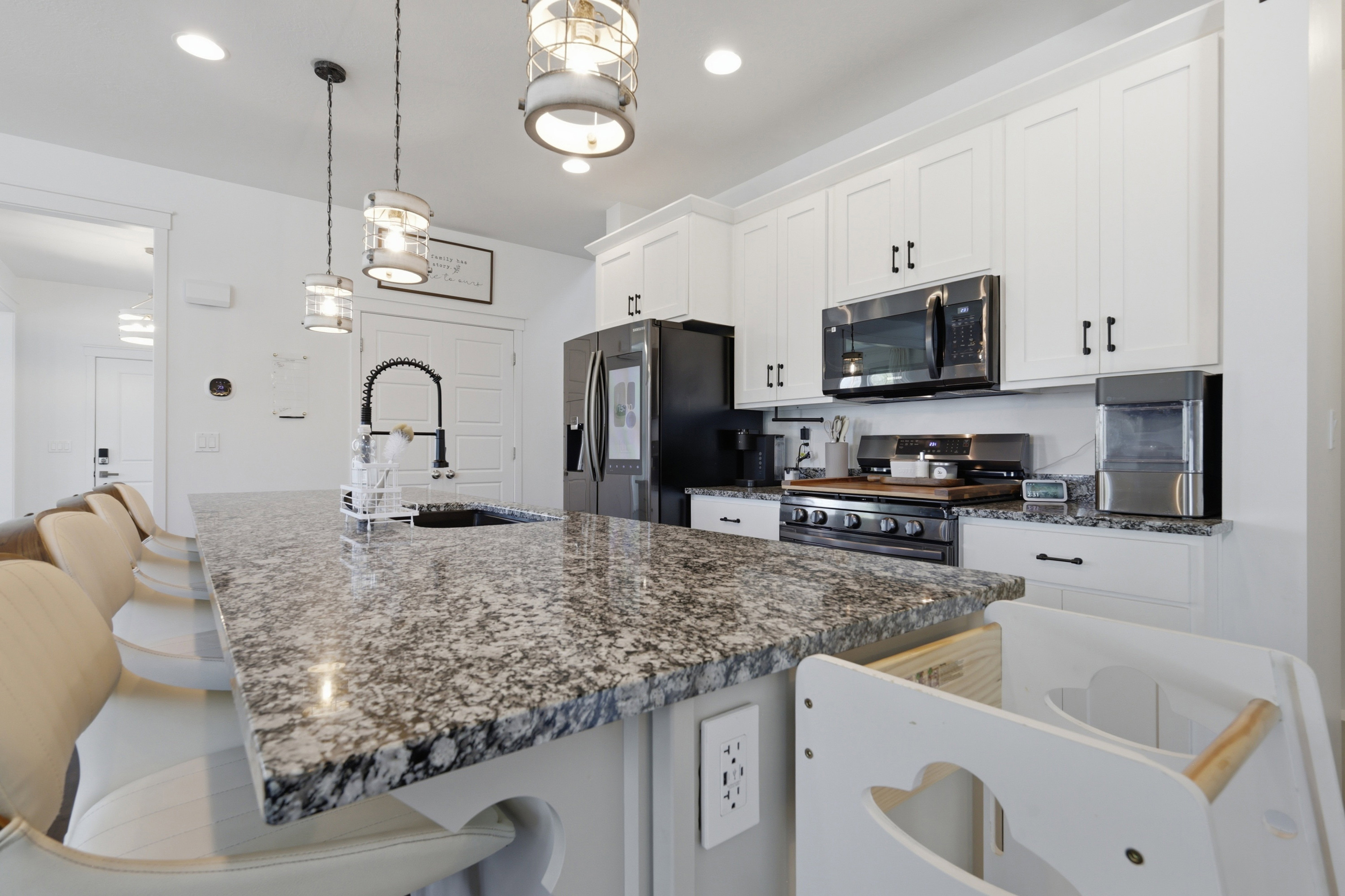 Kitchen featuring a spacious island with a dark speckled countertop, white shaker-style cabinetry with dark hardware, black stainless steel appliances, and ceiling-mounted pendant lighting