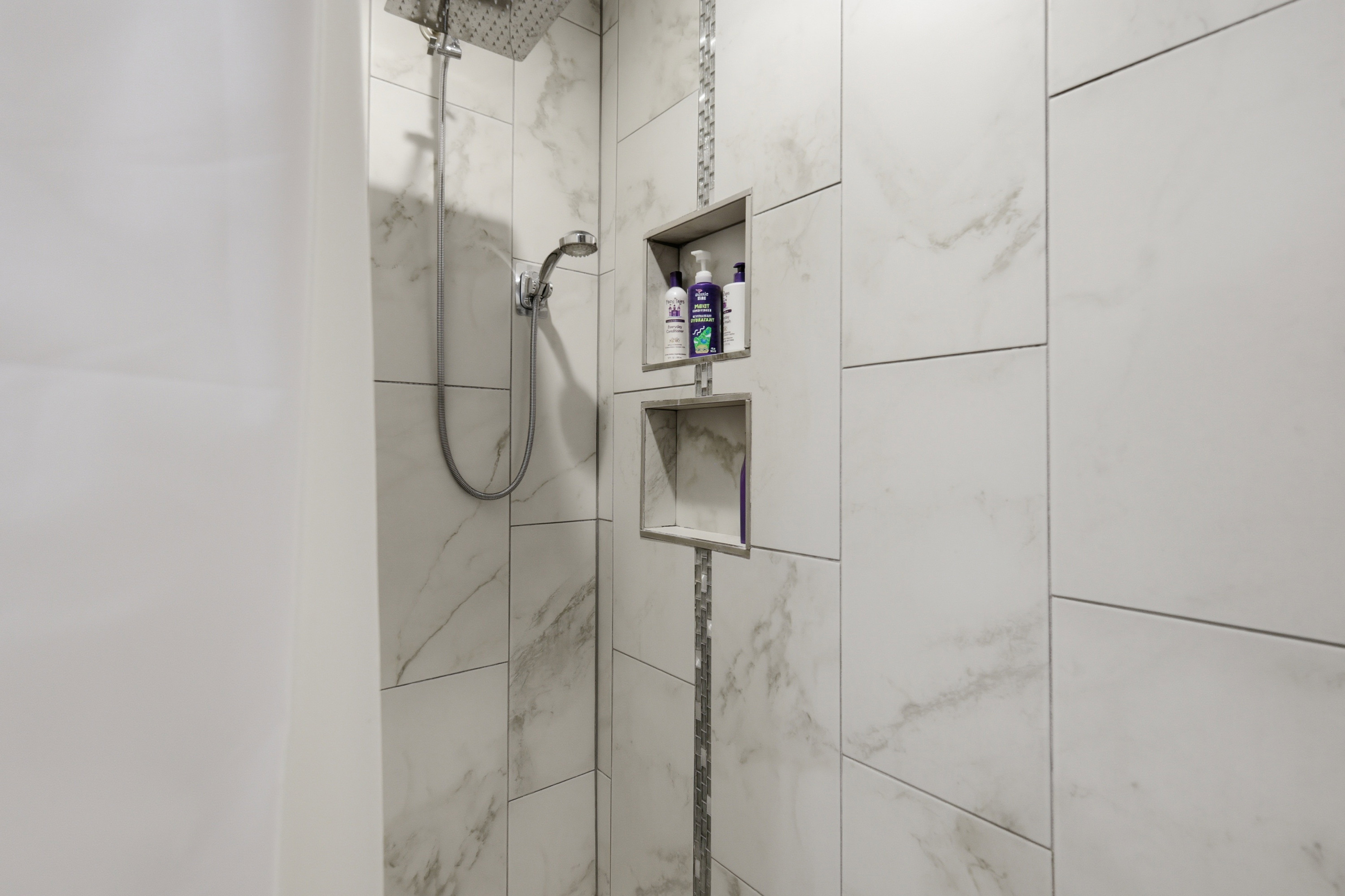 Shower with large-format white tile, chrome fixtures including a rain showerhead and handheld sprayer, and built-in recessed shelving