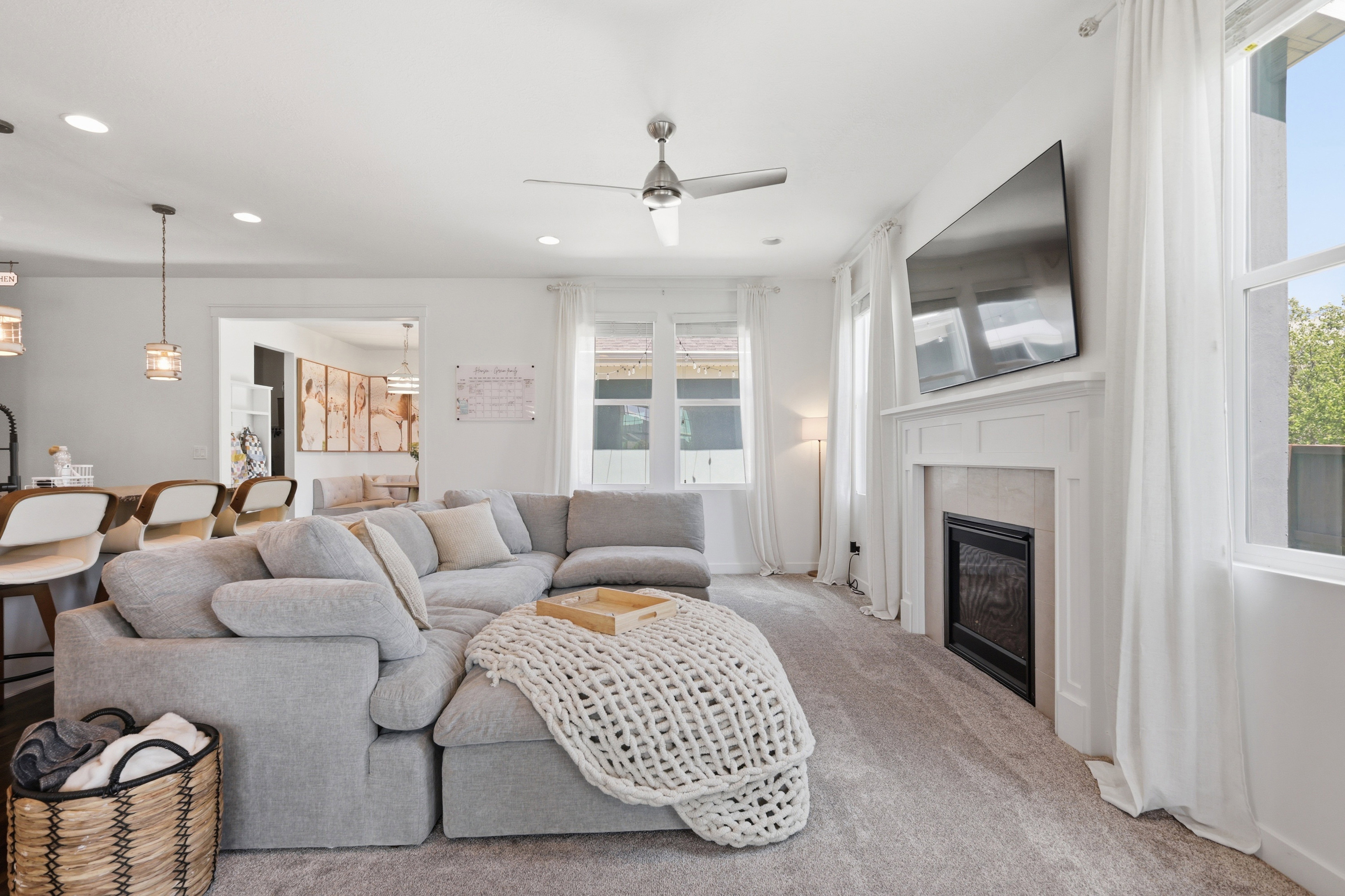 Light-filled living room featuring a recessed fireplace with a white mantel, ceiling fan, and large windows with white drapes