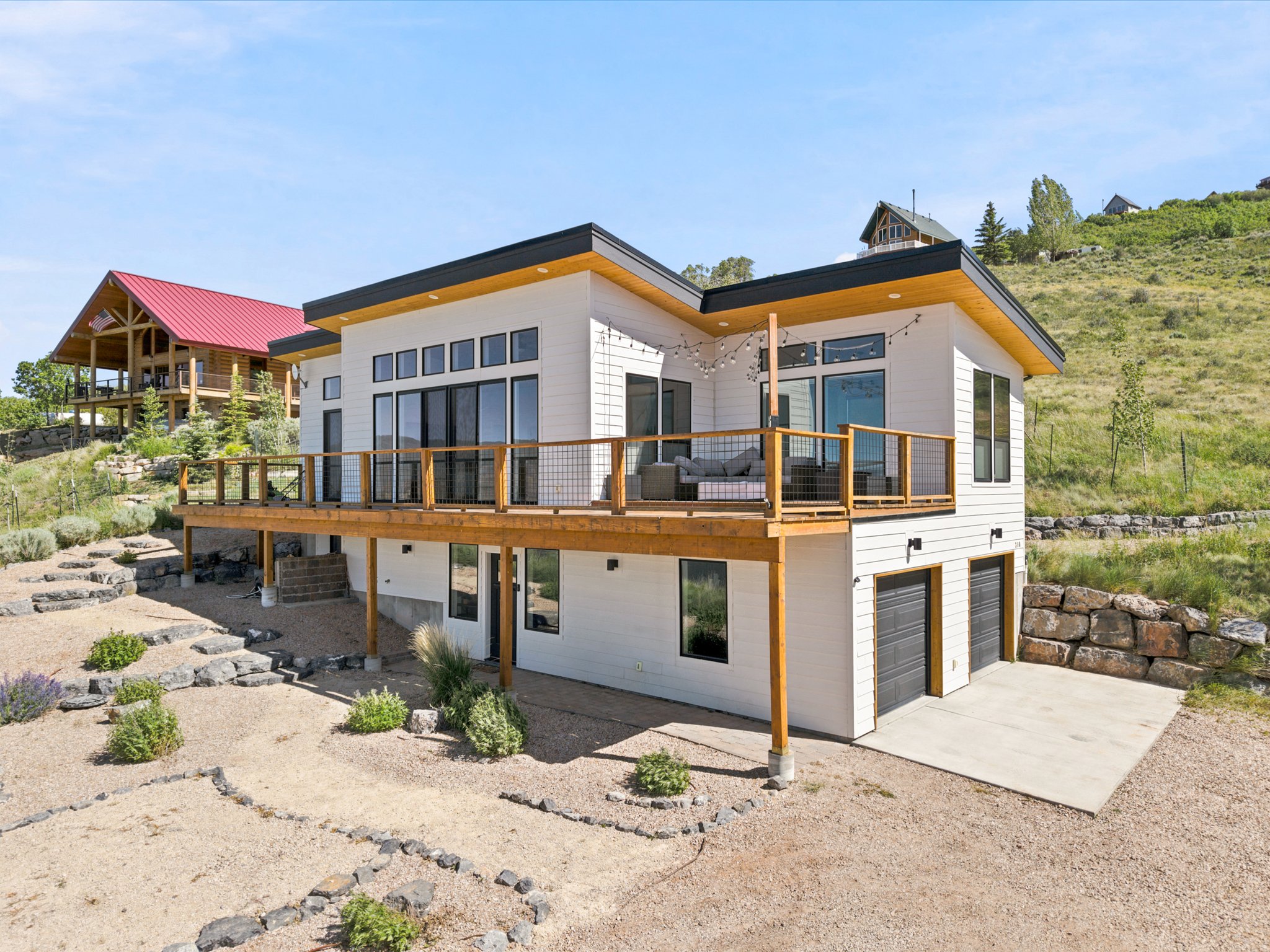 Modern white exterior with a large wood-finish deck and cable railings, featuring a two-car garage with contemporary doors and a gravel driveway