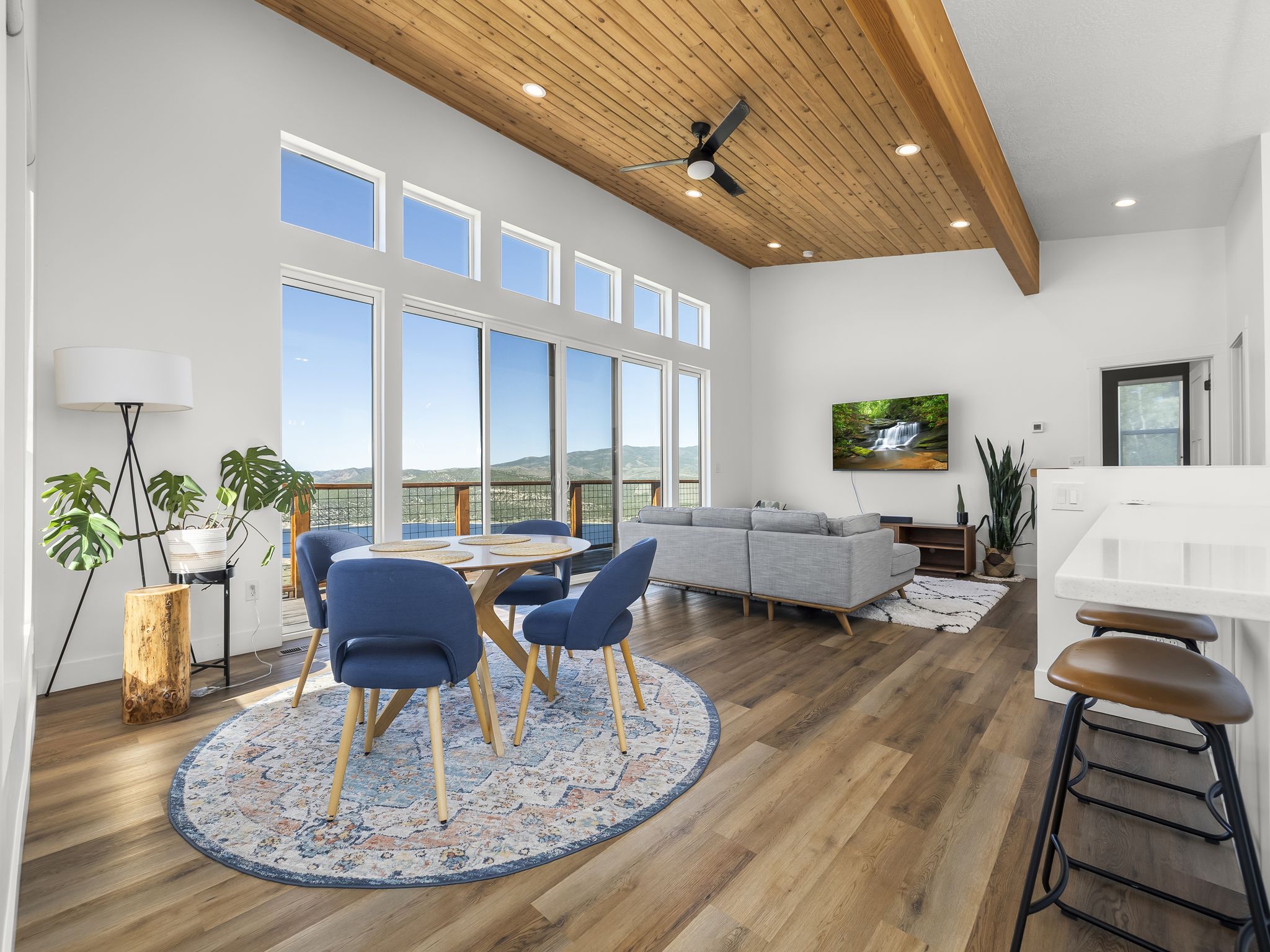 Expansive living area featuring soaring vaulted ceilings with wood paneling and exposed beams, large format windows, wood-finish flooring, and a built-in kitchen island with a white countertop