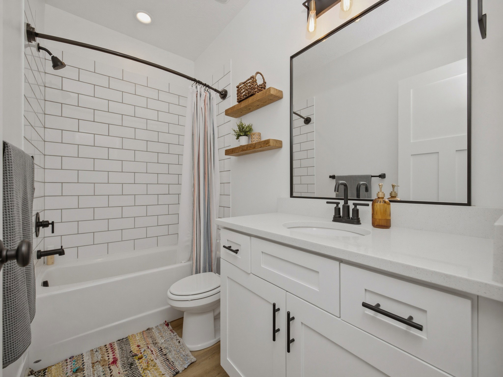 Bathroom featuring a white subway tile shower surround, dark bronze fixtures, a white vanity with a light-toned countertop, a rectangular mirror, and wood-finish flooring