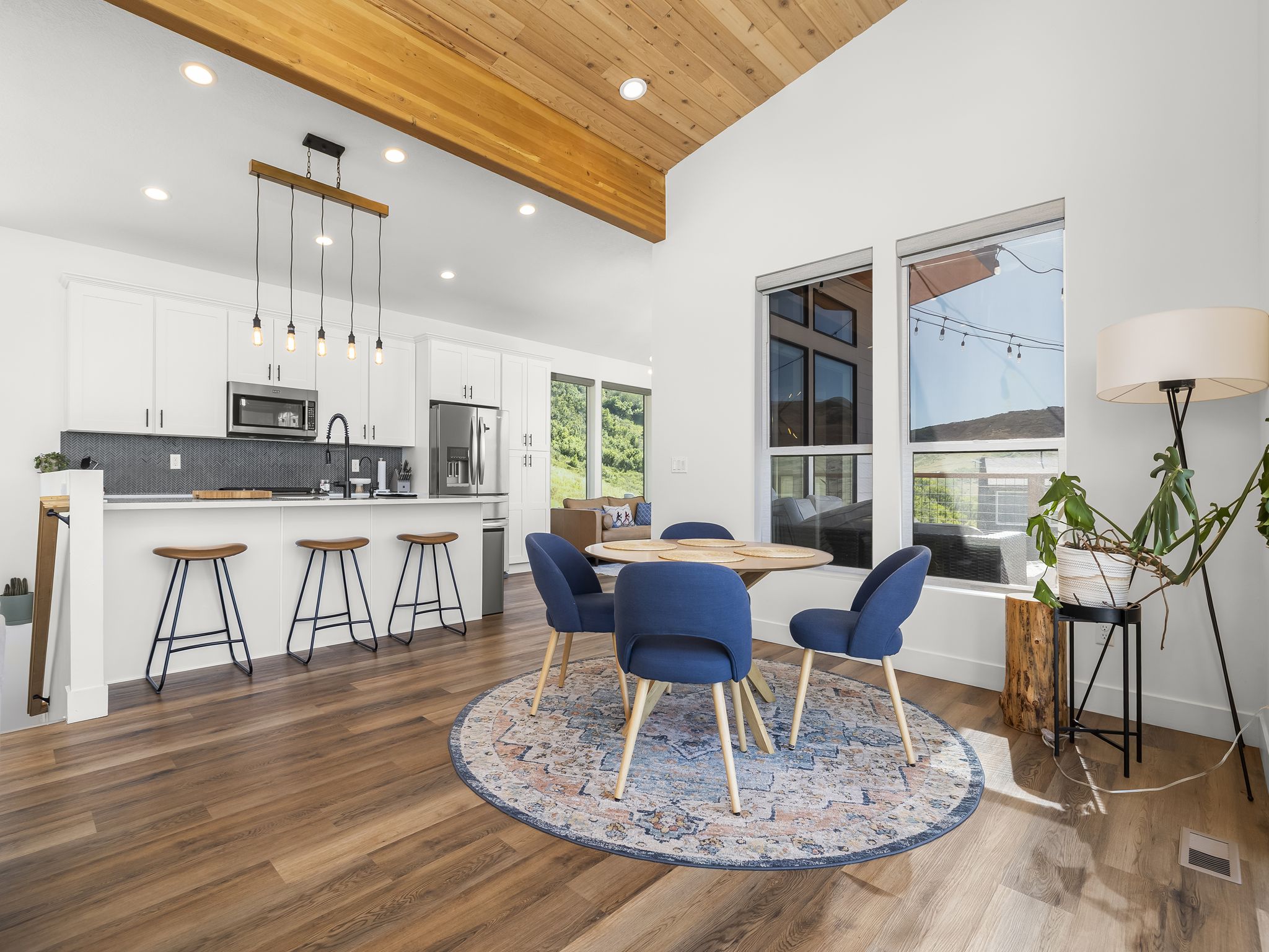 Vaulted wood-paneled ceiling with exposed wood beam, contemporary kitchen with white cabinetry and stainless steel appliances, wood-finish flooring, and multiple large windows