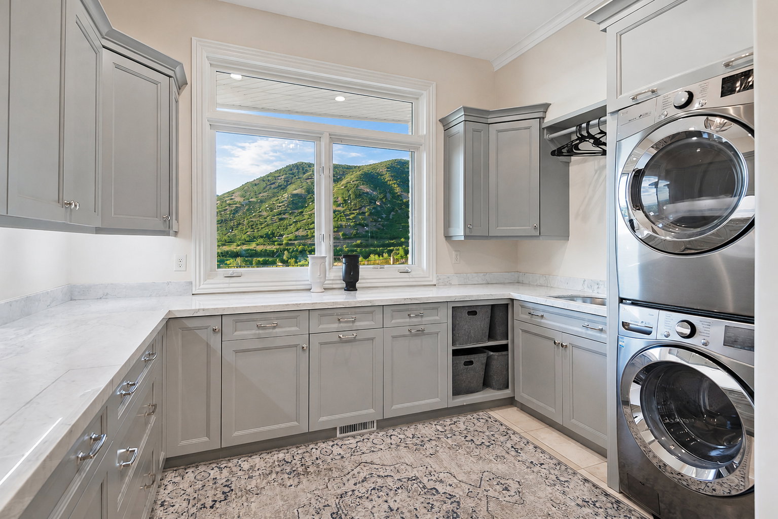 Bright utility room featuring ample light gray cabinetry, white countertops, built-in shelving, a stainless steel stacked washer and dryer, and a large window overlooking a verdant hillside