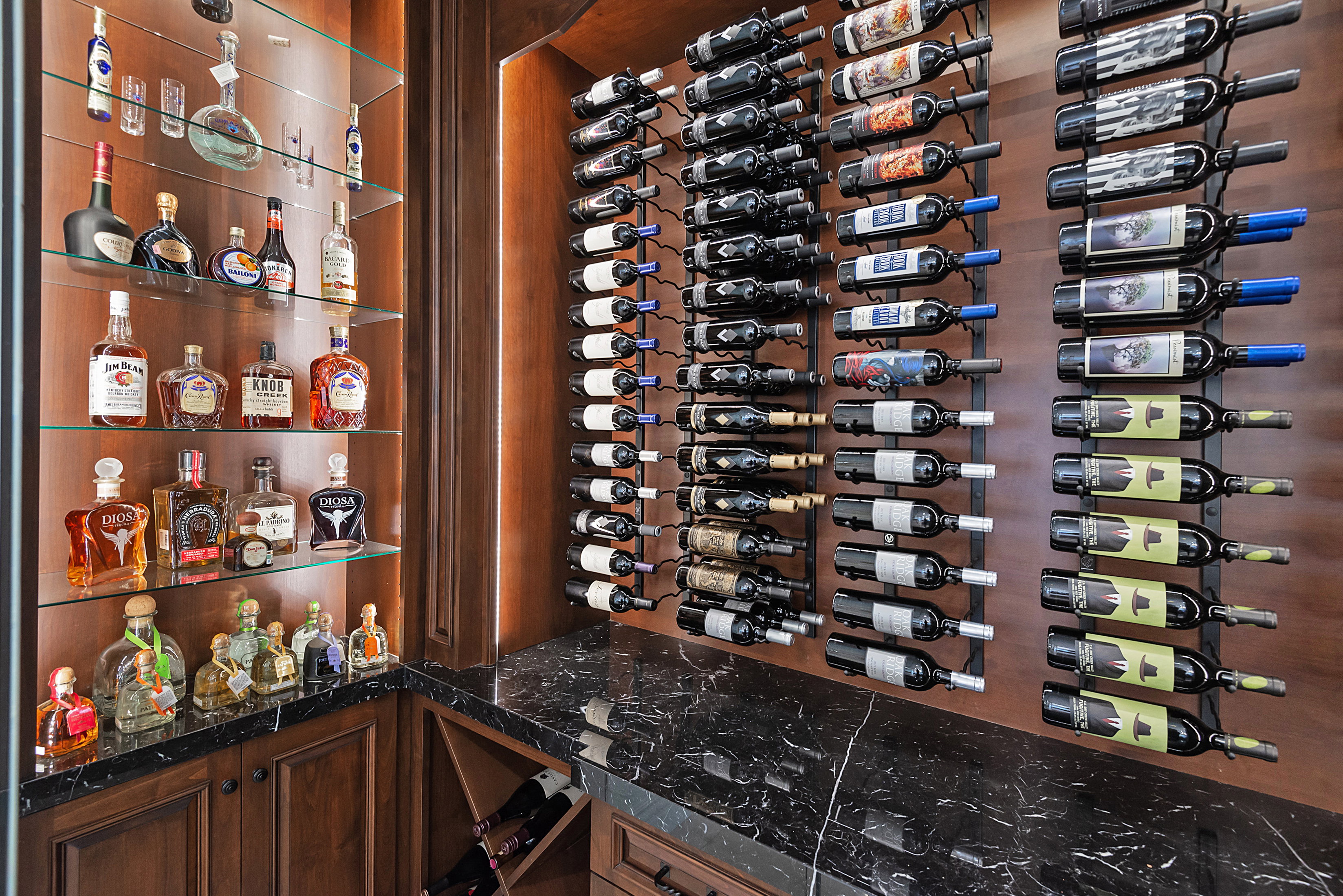 Custom wine storage room featuring warm wood-finish walls, extensive bottle racking, a built-in display cabinet with glass shelving, integrated lighting, and a dark stone-finish countertop