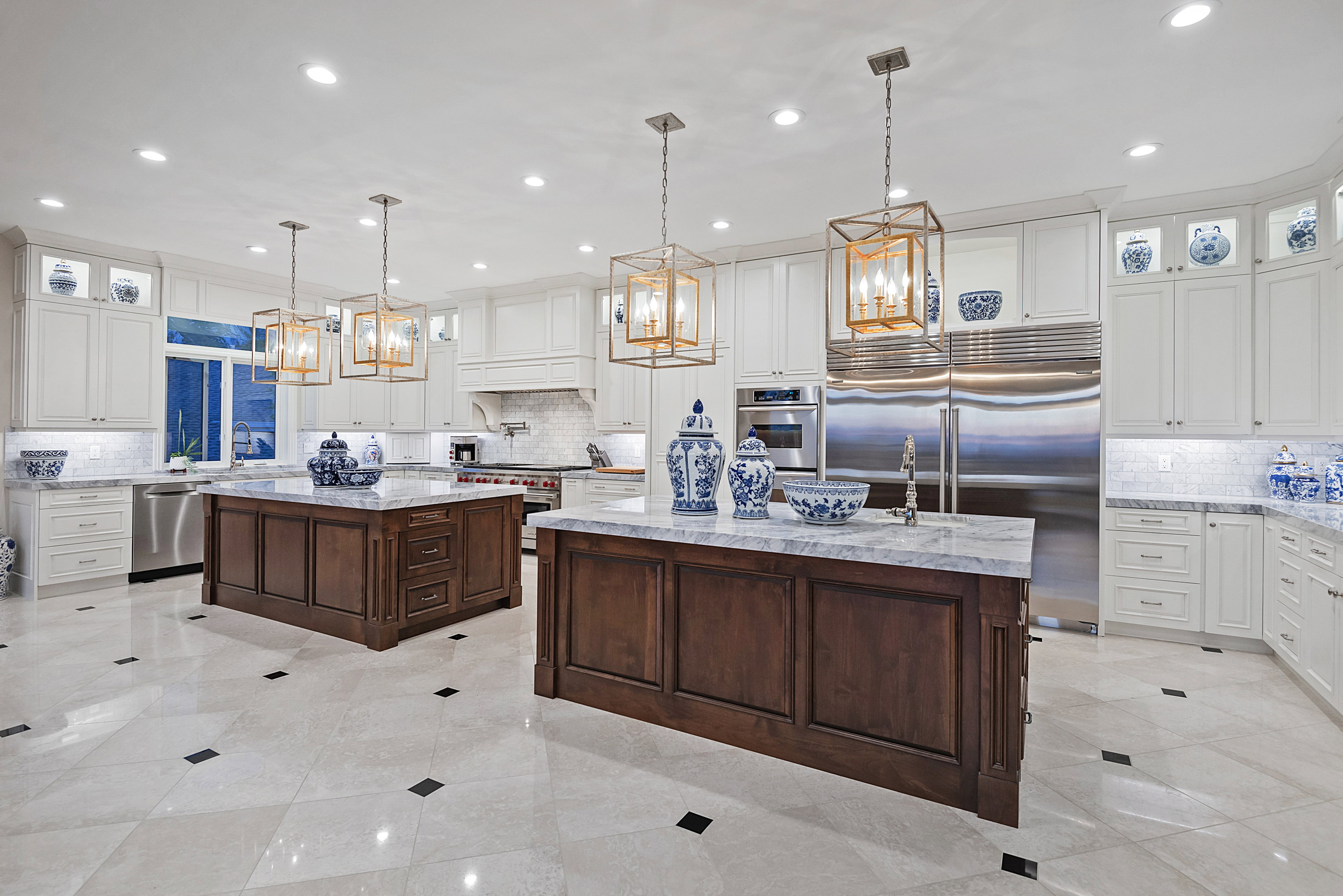 Gourmet kitchen featuring two central islands with stone countertops and wood-panel cabinetry, stainless steel appliances, white perimeter cabinetry, and recessed lighting
